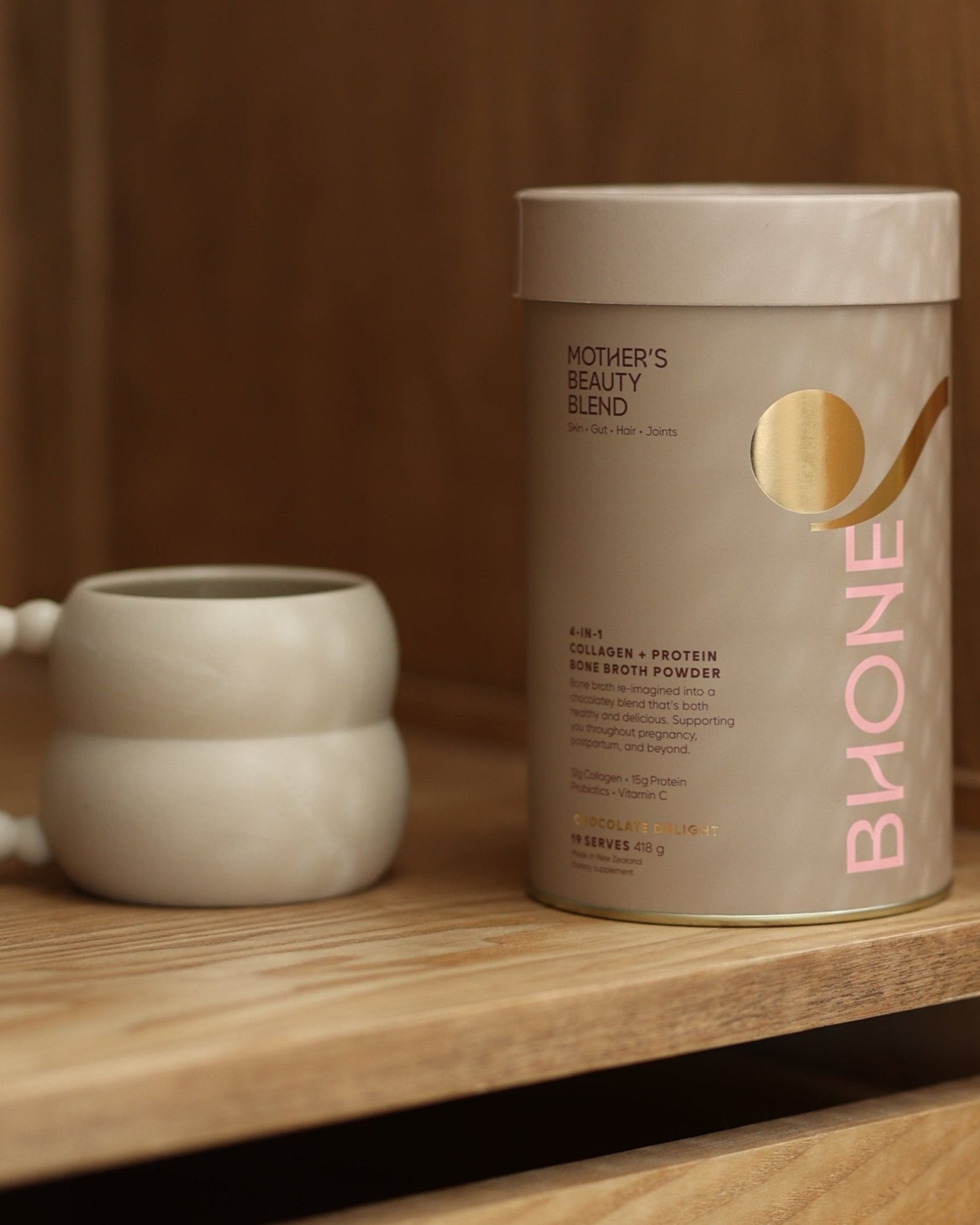 A container of Bhone's Mother's Beauty Blend in Chocolate Haven, a nutrition drink, is displayed next to a mug on a wooden surface.